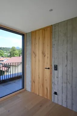 Moderne Holztür in Betonwand neben Fenster mit Blick auf Dächer und bewaldete Hügel