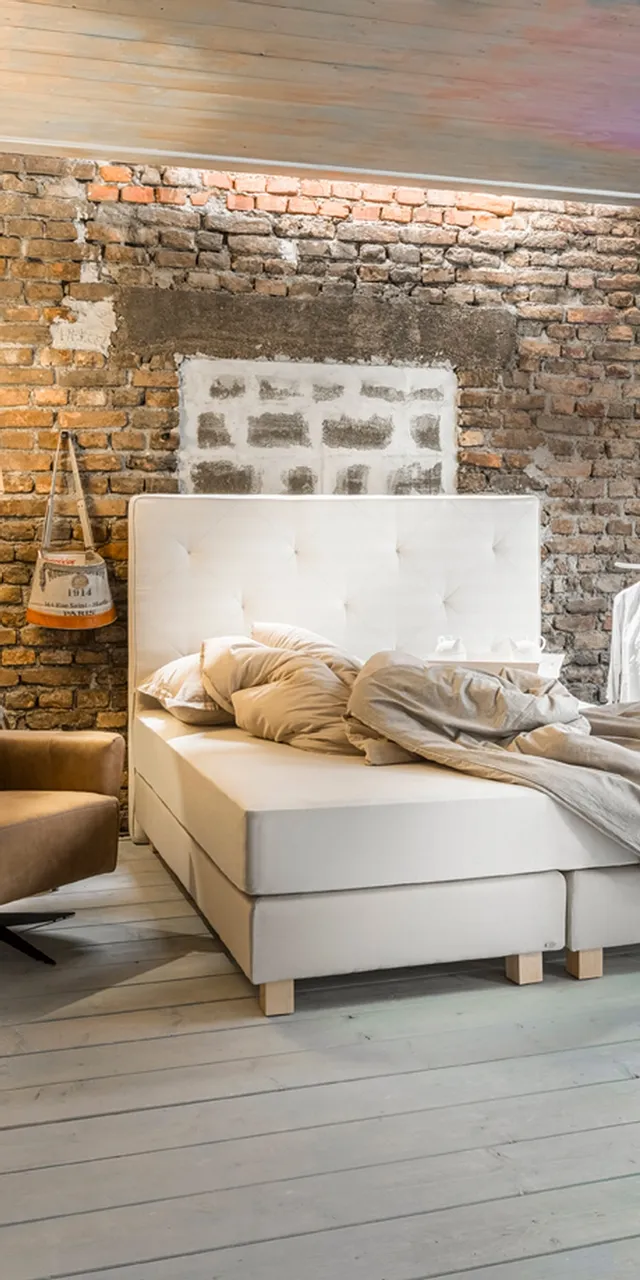 Modernes Schlafzimmer mit Boxspringbett vor rustikaler Backsteinwand und gemütlichem Sessel