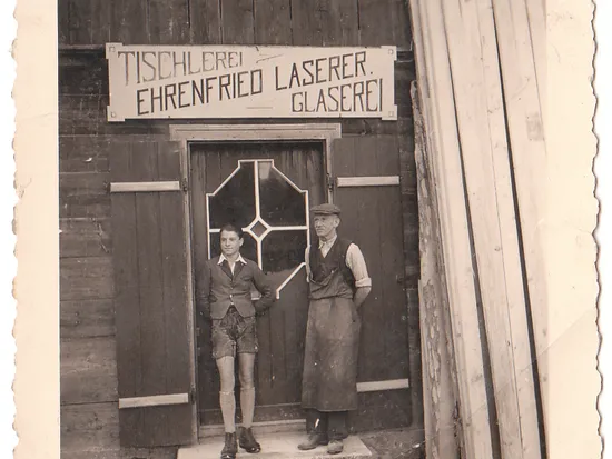Zwei Männer vor einer Tischlerei mit Schild 'Tischlerei Ehrenfried Laserer Glaserei'